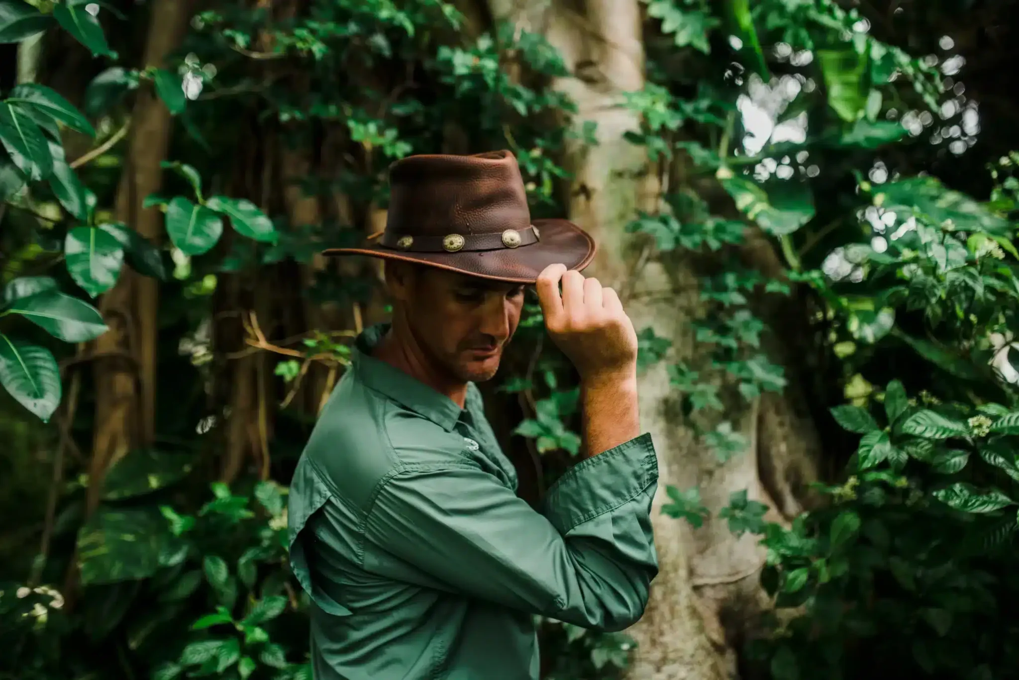 Man in hat tipping it while standing in lush greenery.